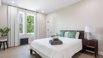 Quarto com cama grande, mesa de cabeceira, luminária, planta em vaso e janela com cortinas.