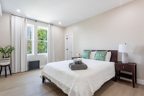Quarto com cama grande, mesa de cabeceira, luminária, planta em vaso e janela com cortinas.