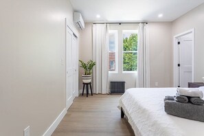 A bedroom with a bed, a radiator, a window with curtains, a potted plant, and a door.