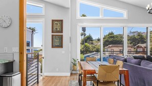 A modern living room with a dining area, large windows, and a view of a residential area. - Capitola Village Luxury 4BR | Hot Tub & Parking (Capitola)