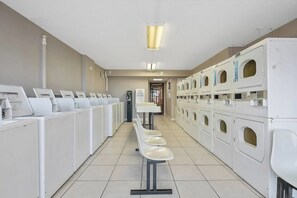 A laundry facility with rows of washing machines and dryers, a seating area with chairs, and a sign indicating the availability of softeners. - Cozy 1-bedroom condo in charming Myrtle Beach with AC and gym (Myrtle Beach)