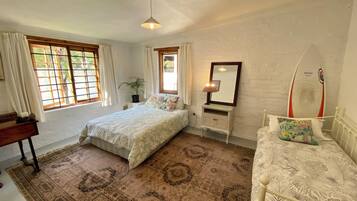 A bedroom with a bed, bedside table, lamp, mirror, surfboard decoration, and a window with curtains.