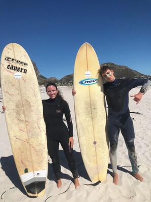 Two individuals holding surfboards on a sandy beach with a clear sky and mountains in the background. - Room-shared Dolphin Dorm Room (Hermanus)