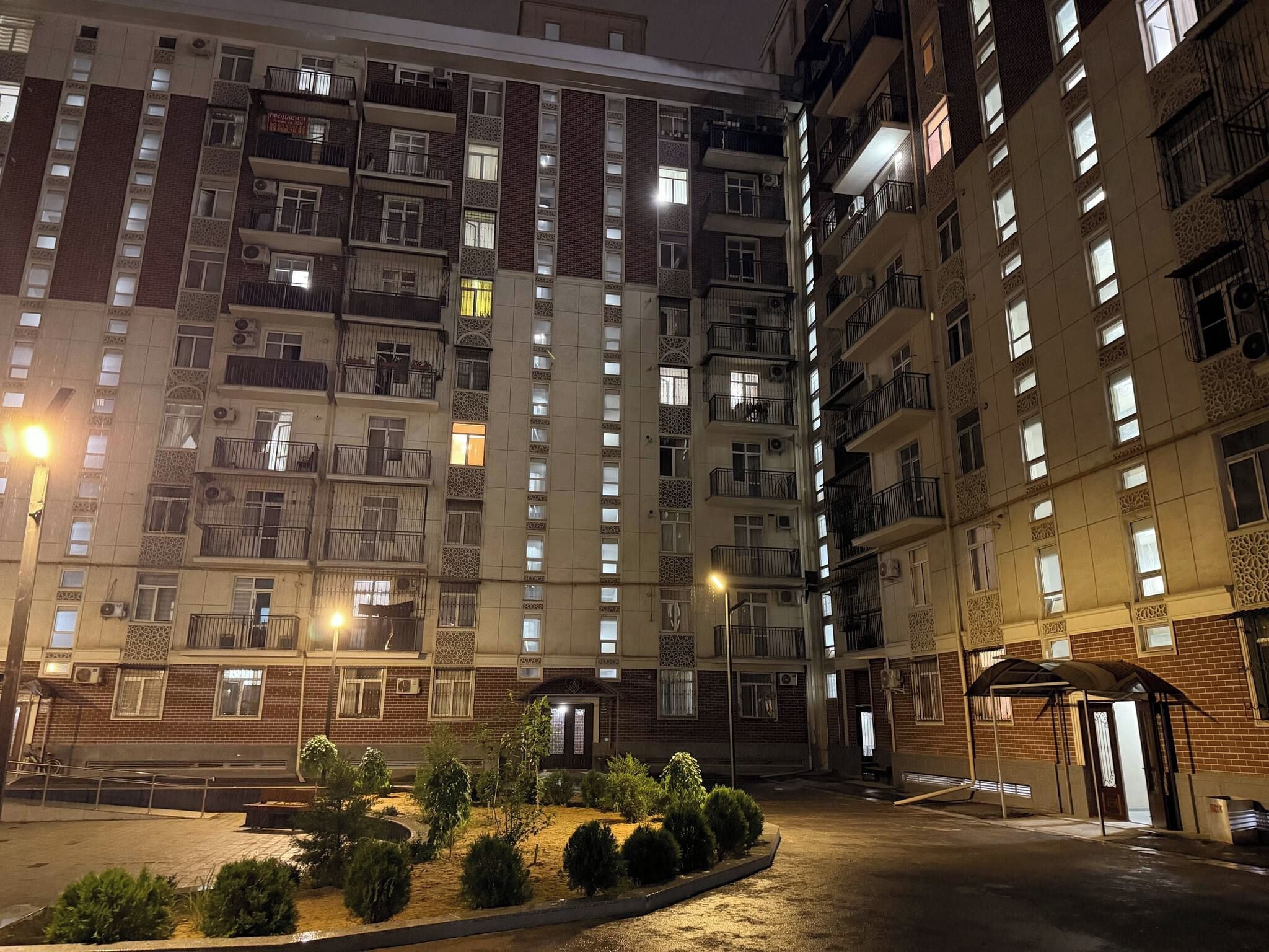 A multi-story residential building with balconies, a gated entrance, and a small landscaped area with shrubs.