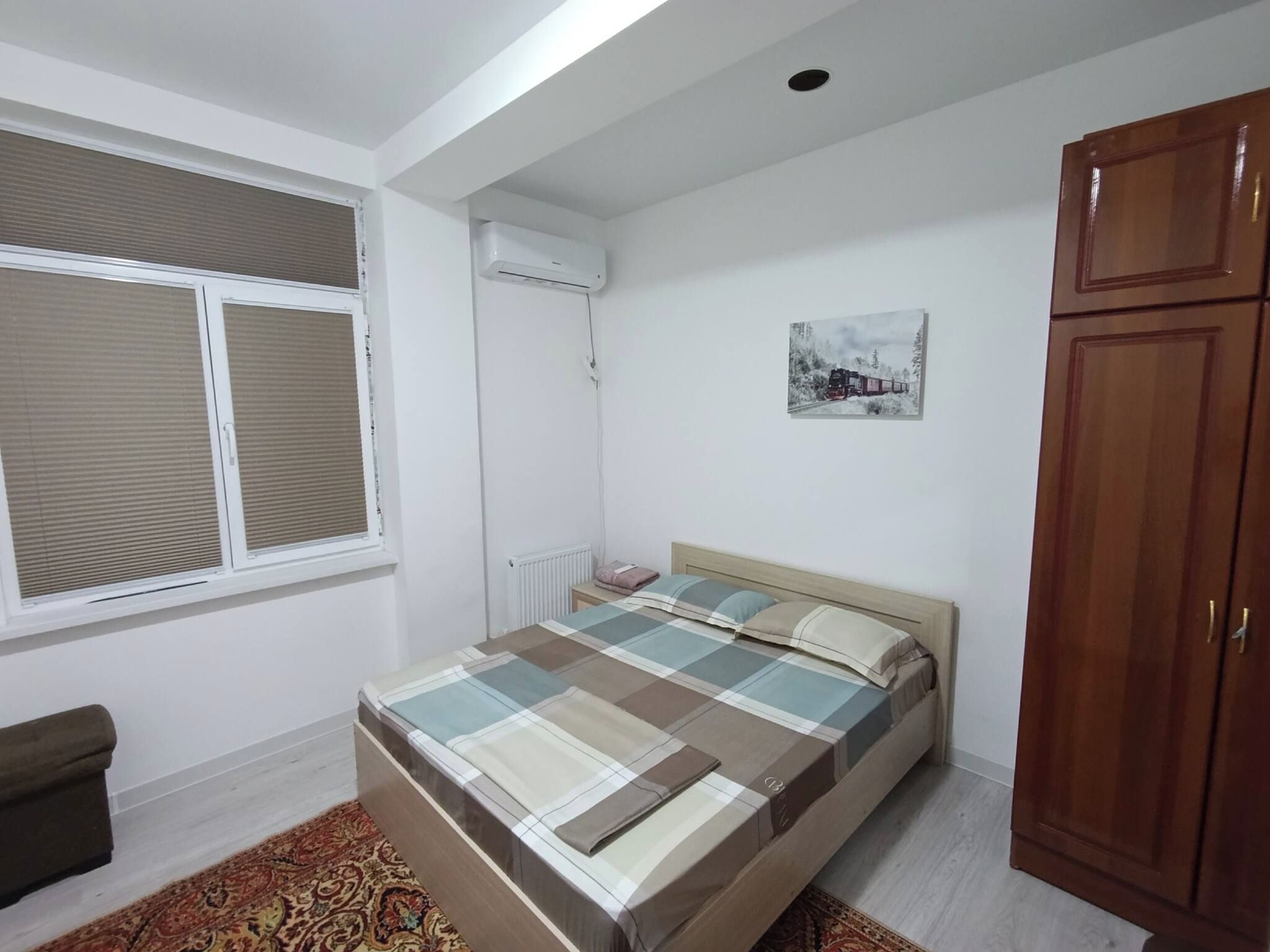 A bedroom with a bed, a window with blinds, a radiator, a wall-mounted air conditioner, a framed picture, and a wardrobe.
