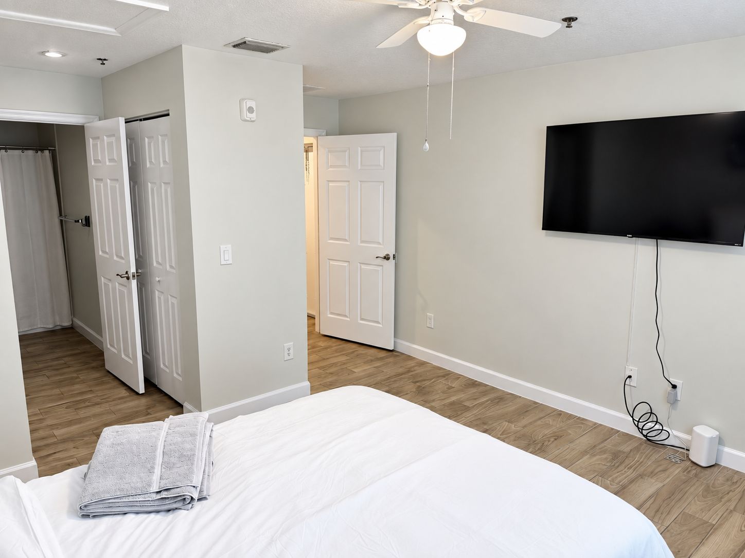 A bedroom with a bed, a ceiling fan, a TV mounted on the wall, and a door leading to another room.