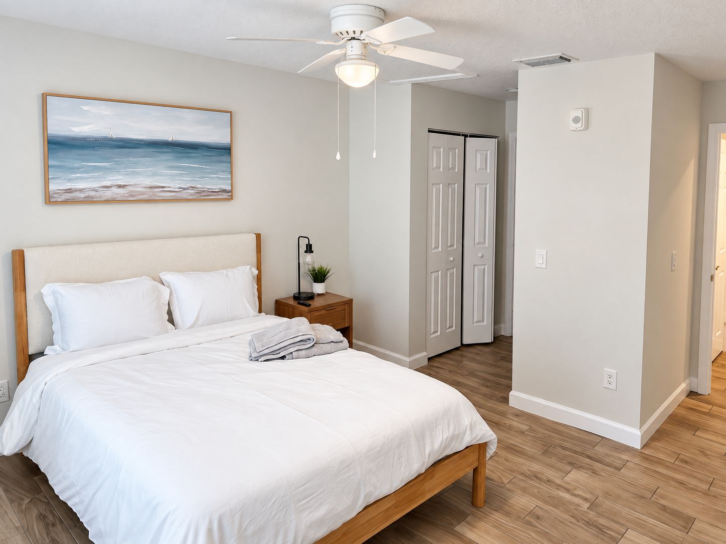 A bedroom with a bed, a nightstand with a lamp, a ceiling fan, and a door leading to another room.