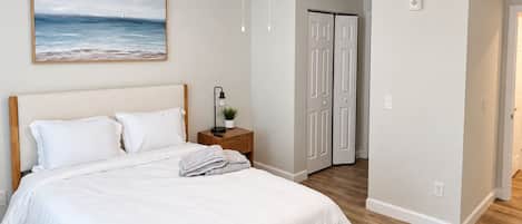 A bedroom with a bed, a nightstand with a lamp, a ceiling fan, and a door leading to another room.