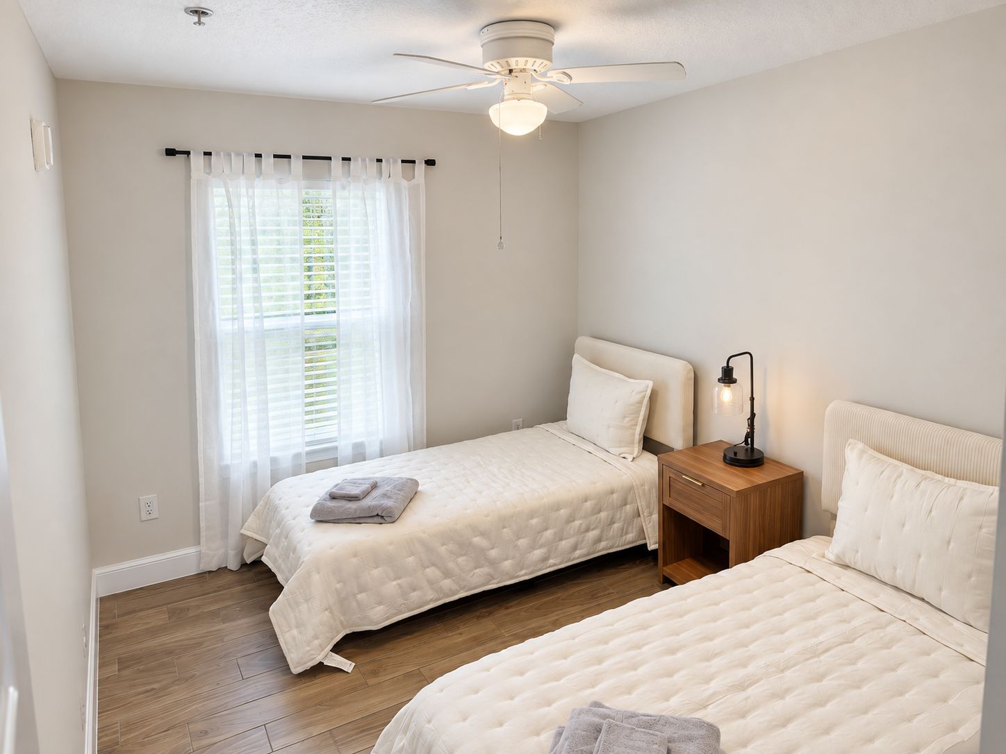 A bedroom with two beds, a ceiling fan, a nightstand with a lamp, and a window with curtains.