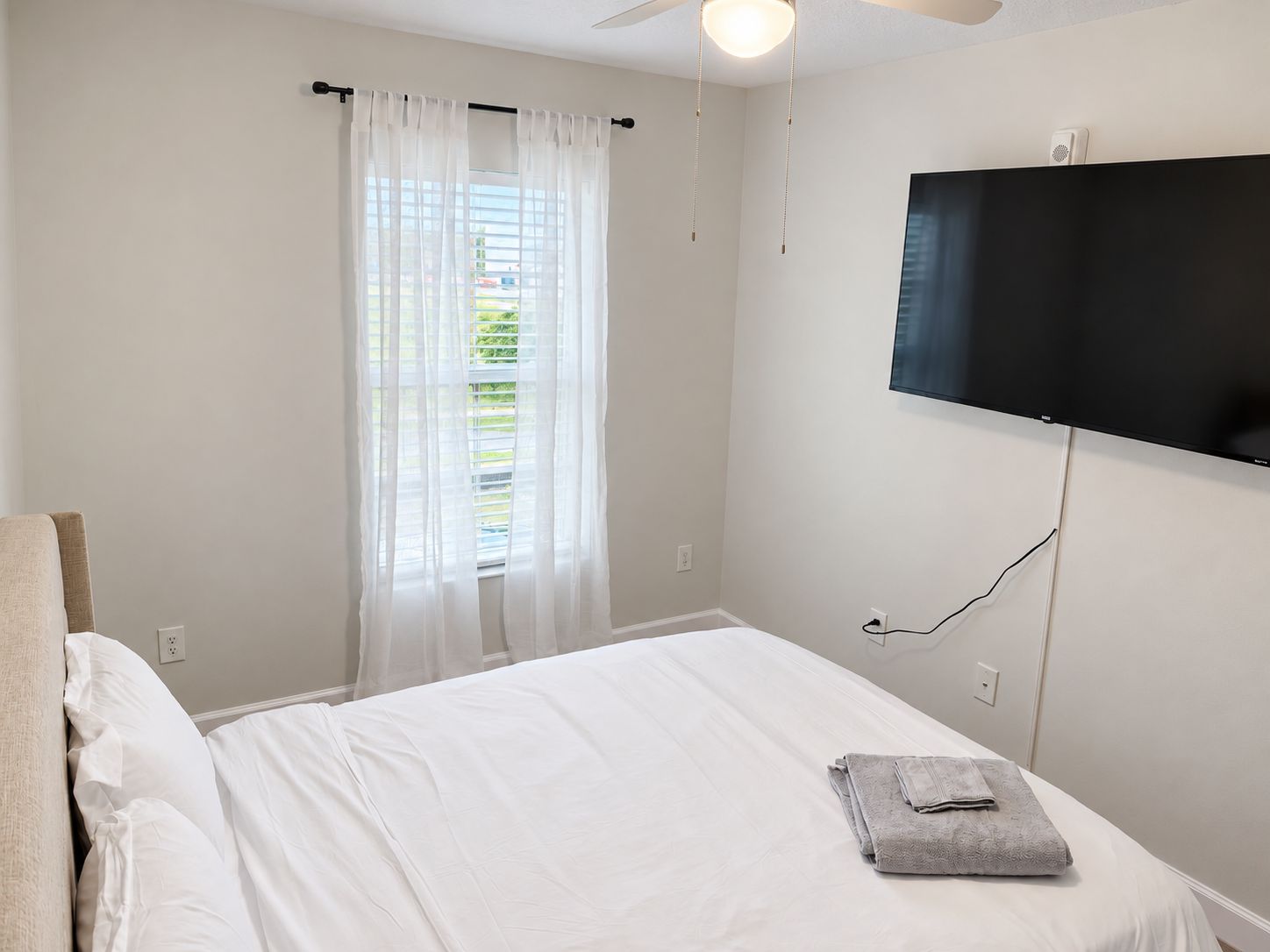 Une chambre à coucher avec un lit, un téléviseur fixé au mur, une fenêtre avec des rideaux légers et un ventilateur de plafond.
