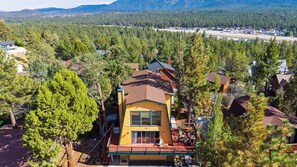 A two-story house with a red roof, surrounded by dense forest, and a clear view of a distant town. - Bear Pine Lodge by Avantstay Hot Tub + Firepit + Arcade Mountain Views Mins to Slopes + Lake (Big Bear City)