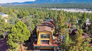 A two-story house with a red roof, surrounded by dense forest, and a clear view of a distant town. - Bear Pine Lodge by Avantstay Hot Tub + Firepit + Arcade Mountain Views Mins to Slopes + Lake (Big Bear City)