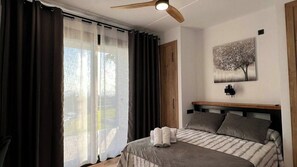 Ein Schlafzimmer mit einem Bett, einem Deckenventilator, einem Fenster mit Vorhängen und einer Wandleuchte.