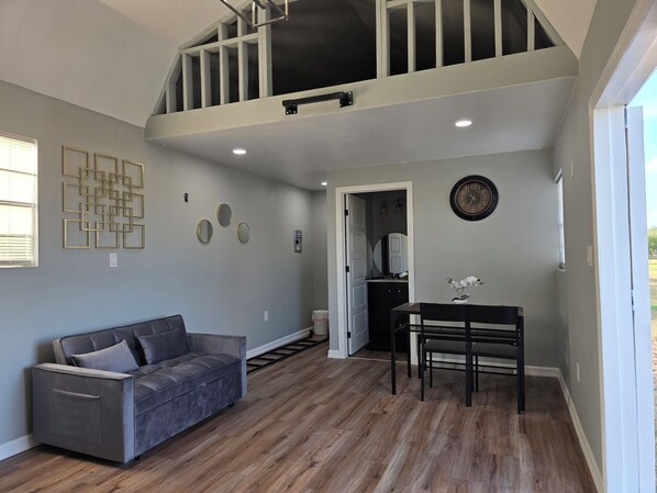 A spacious living area with a grey sofa, a dining table with chairs, a staircase, and a large wall clock.