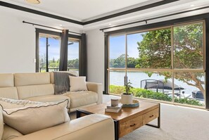 A modern living room with a large window, a beige sofa, and a coffee table. - Waterfront 5BR + Pool | Kawana Island | Sleeps 10 (Parrearra)