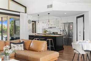 A modern kitchen with white cabinetry, a central island, and a dining area with a white table and chairs. - Waterfront 5BR + Pool | Kawana Island | Sleeps 10 (Parrearra)
