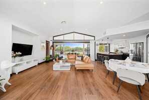 A spacious living room with a large flat-screen TV, a leather sofa, and a dining area with a table set for two. - Waterfront 5BR + Pool | Kawana Island | Sleeps 10 (Parrearra)