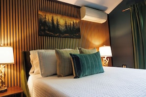 A hotel room with a bed, two bedside tables with lamps, a wall-mounted air conditioner, and a painting of a forest scene. - Cozy Lakefront home in the heart of the White Mountains (Conway)