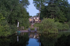 A lakeside cabin with a deck and outdoor seating, surrounded by dense forest. - Cozy Lakefront home in the heart of the White Mountains (Conway)