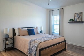 A bedroom with a bed, bedside lamps, a window with curtains, and a wall-mounted shelf with a plant.