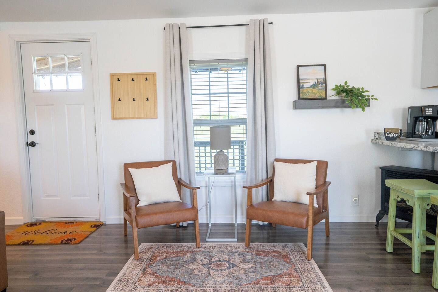 A room with two wooden chairs, a patterned rug, a door, a window with curtains, a coffee station, and a welcome mat.