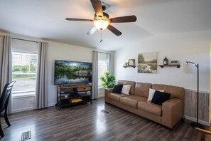 A living room with a brown leather sofa, a flat-screen TV, a ceiling fan, and a large window with blinds.