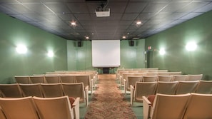 A conference room with rows of chairs facing a large white screen, green walls, and a carpeted floor.
