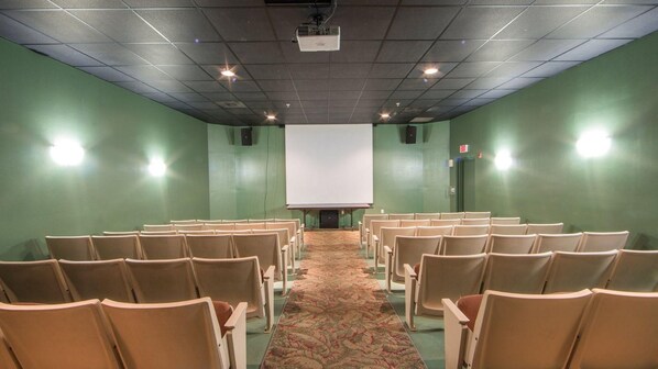 A conference room with rows of chairs facing a large white screen, green walls, and a carpeted floor.