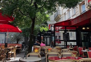 An outdoor cafe with wicker chairs and red umbrellas, a tree in the center, and a building with a sign that reads 'L'Atelier Bulle.' - JUST MOMENTS FROM THE LOUVRE, OPÉRA GARNIER & GRANDS BOULEVARDS (Paris)