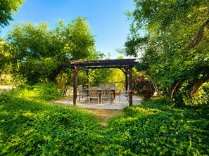 Une pergola en bois, agrémentée d’une table et de chaises, entourée d’une végétation luxuriante et d’arbres.