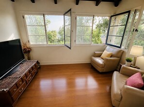 Un salon avec un téléviseur, un meuble en bois, un canapé beige et de grandes fenêtres donnant sur la vue des arbres.