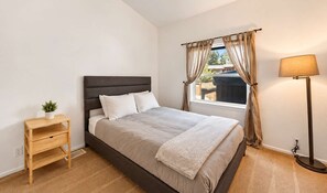 A bedroom with a bed, a bedside table with a plant, a window with curtains, and a floor lamp. - Modern Retreat W Hot Tub & Near Snow Summit (Big Bear Lake)