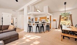A spacious living area with a sofa, a dining table with chairs, a kitchen with bar stools, and a large window with curtains. - Modern Retreat W Hot Tub & Near Snow Summit (Big Bear Lake)