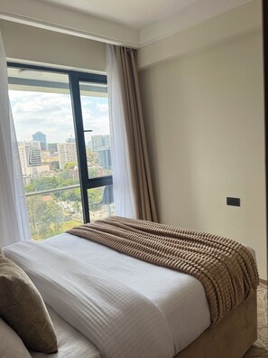 A hotel room with a large window, a bed with a textured blanket, and a city view. - 15th floor | Heart of Westlands | Skyline | Heated pool | Near GTC & Sarit (Nairobi)