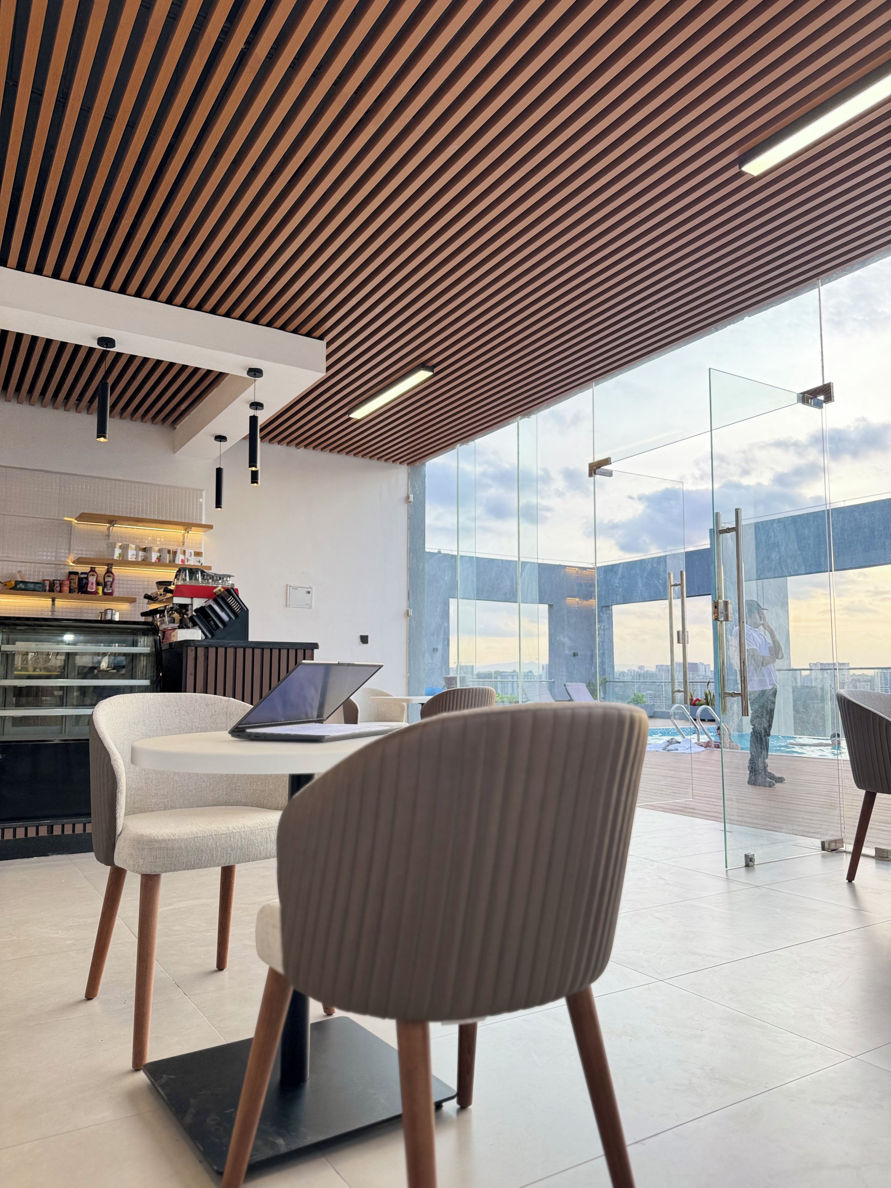 Un interno moderno con soffitto in legno, una parete vetrata con vista su una piscina e un’area salotto con un laptop su un tavolo.