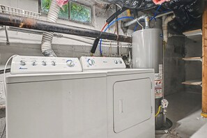 Une machine à laver Amana blanche, dotée de boutons de commande, un chauffe-eau et des tuyaux apparents, dans une pièce utilitaire.