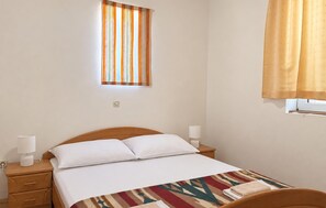 Ein Einzelbett mit weißer Bettwäsche, ein Nachttisch mit Lampe, ein Fenster mit gestreifter Gardine und eine schlichte Wand.