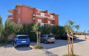 A multi-story building with a red facade, parked cars, and a gravel driveway. - 2 bedroom nice apartment in Razanac (Razanac)