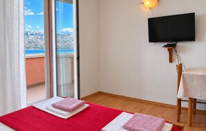 Quarto de hotel com televisão, cama com roupa de cama vermelha e branca, uma cadeira de madeira e vista para uma paisagem montanhosa através de uma porta deslizante.