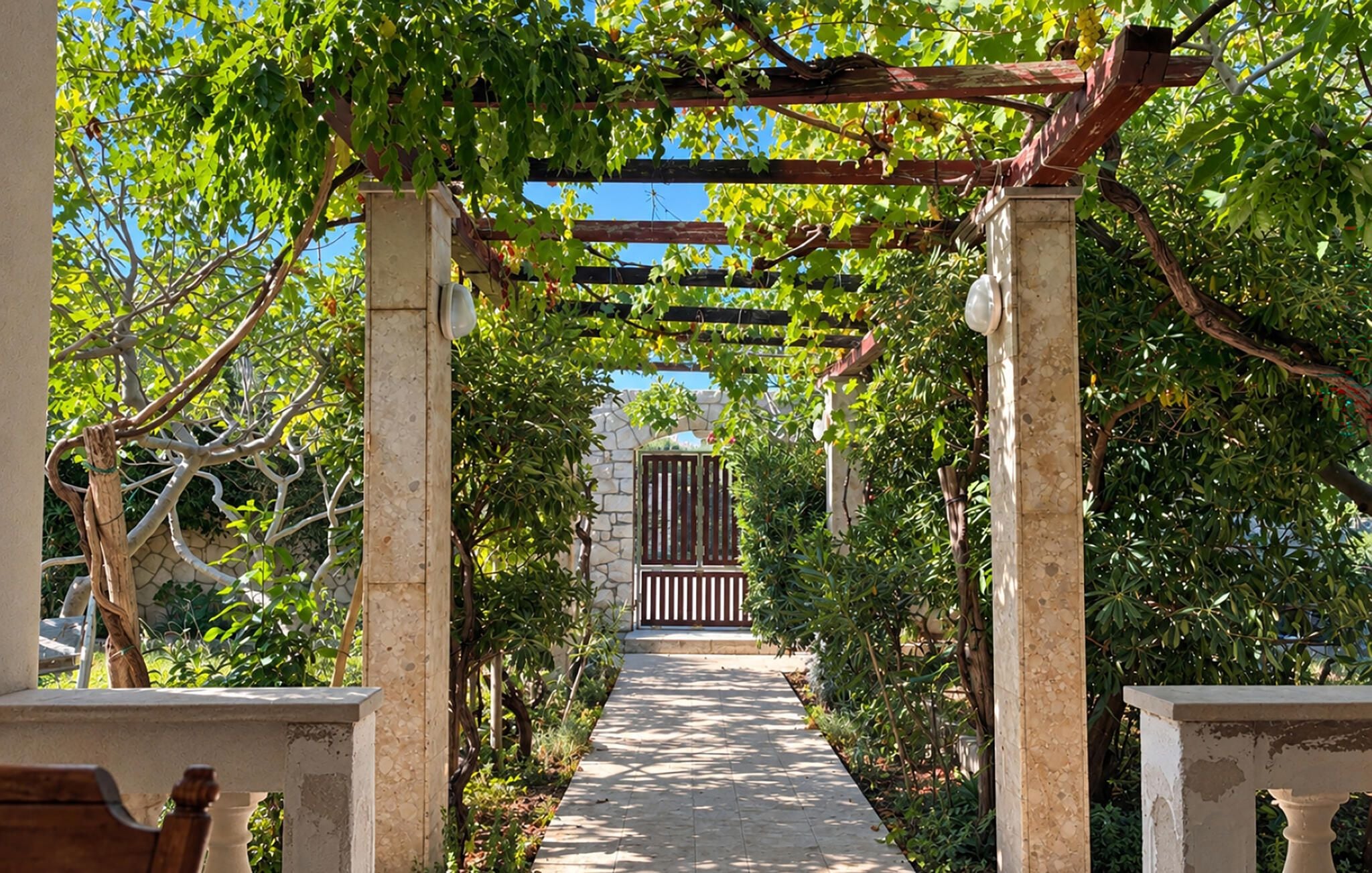 A pathway leading to a gated entrance, flanked by stone pillars and covered by a wooden pergola with vines.