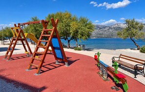A playground with swings and a slide, a bench, and a view of a body of water with mountains in the background. - 1 bedroom lovely apartment in Razanac (Razanac)