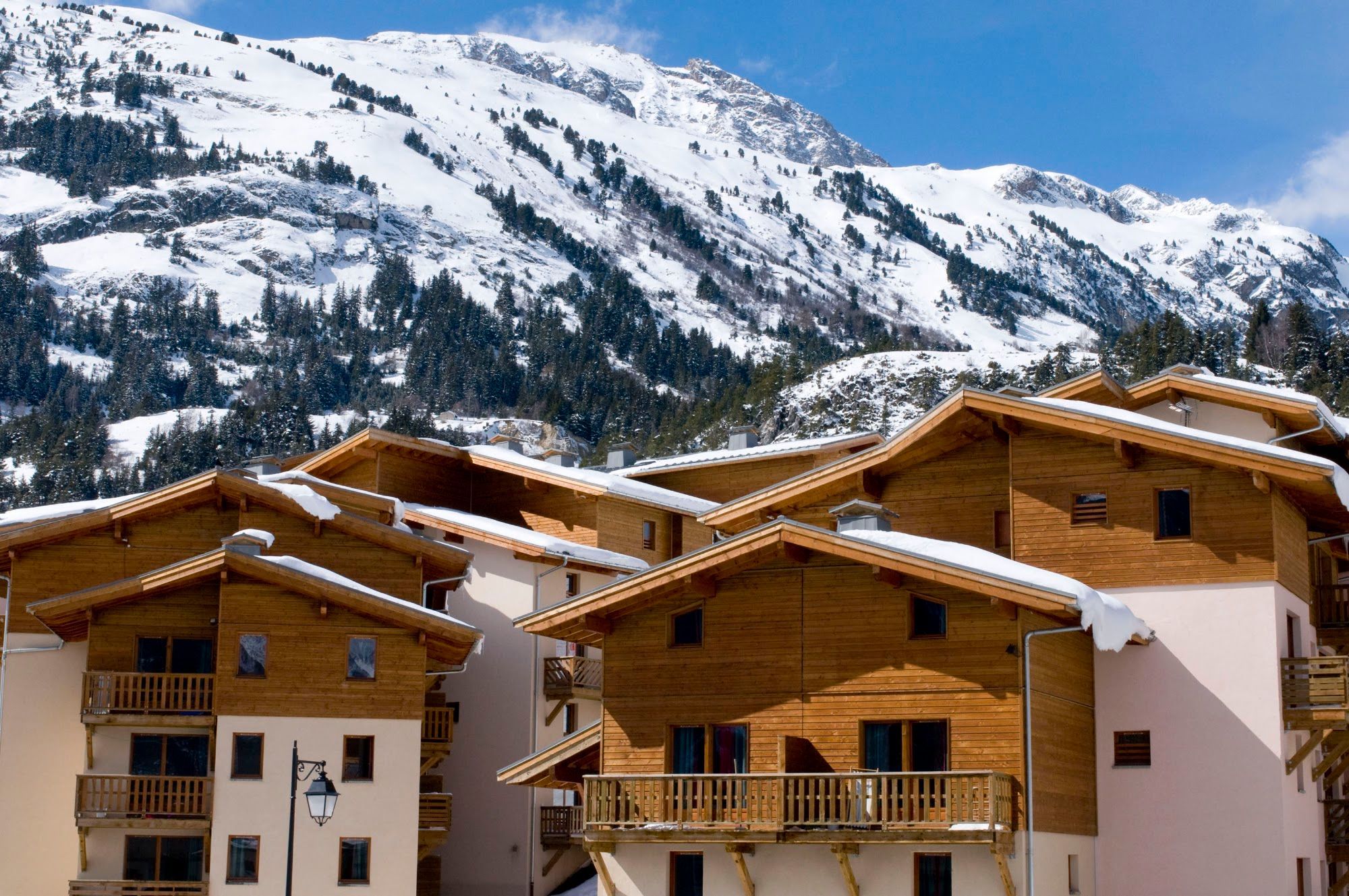 A row of wooden chalet-style buildings with balconies, set against a backdrop of snow-covered mountains.