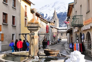 A historic town square with a traditional water pump, cobblestone streets, and a mountain backdrop. - Au pied des pistes | Piscine intérieure chauffée (Aussois)