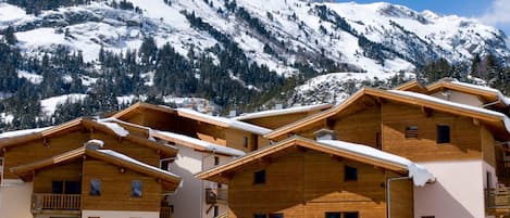 A row of wooden chalet-style buildings with balconies, set against a backdrop of snow-covered mountains.