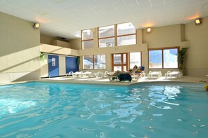 An indoor swimming pool with multiple lounge chairs and a view of a building through large windows. - Au pied des pistes | Piscine chauffée | Balcon (Aussois)