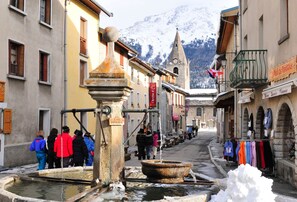 A historic town square with a traditional water pump, cobblestone streets, and a mountain backdrop. - Au pied des pistes | Piscine chauffée | Balcon (Aussois)