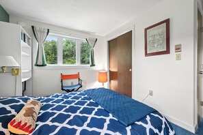 Ein Schlafzimmer mit einem Bett, einem Sessel, einem Fenster mit Vorhängen, einer Lampe und einer Tür.