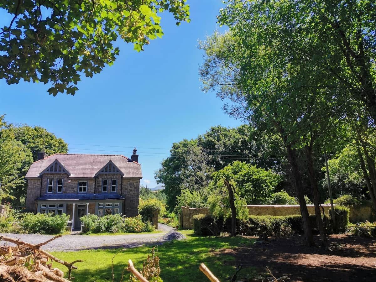 A two-story stone house with a gabled roof, surrounded by lush greenery and a clear blue sky.