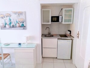 Een compacte keuken met een magnetron, spoelbak en een kleine koelkast. Er staat een eettafel met stoelen en er hangt een schilderij aan de muur.