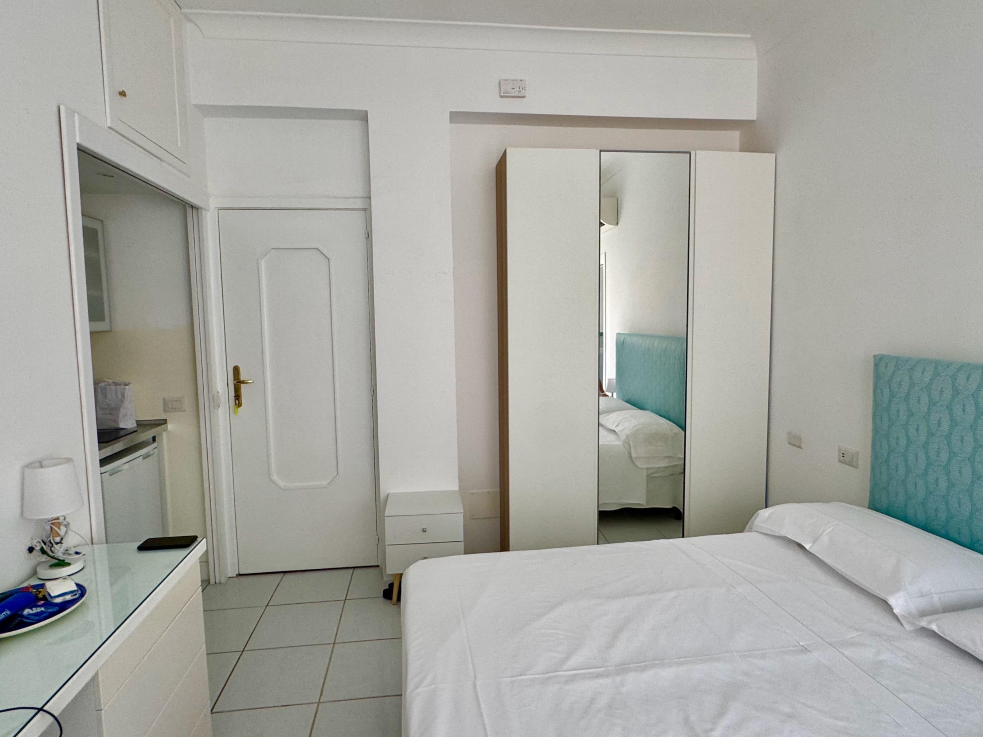 Une chambre d’hôtel comprenant un lit, une table de chevet, un miroir, une armoire et un petit bureau avec une lampe.